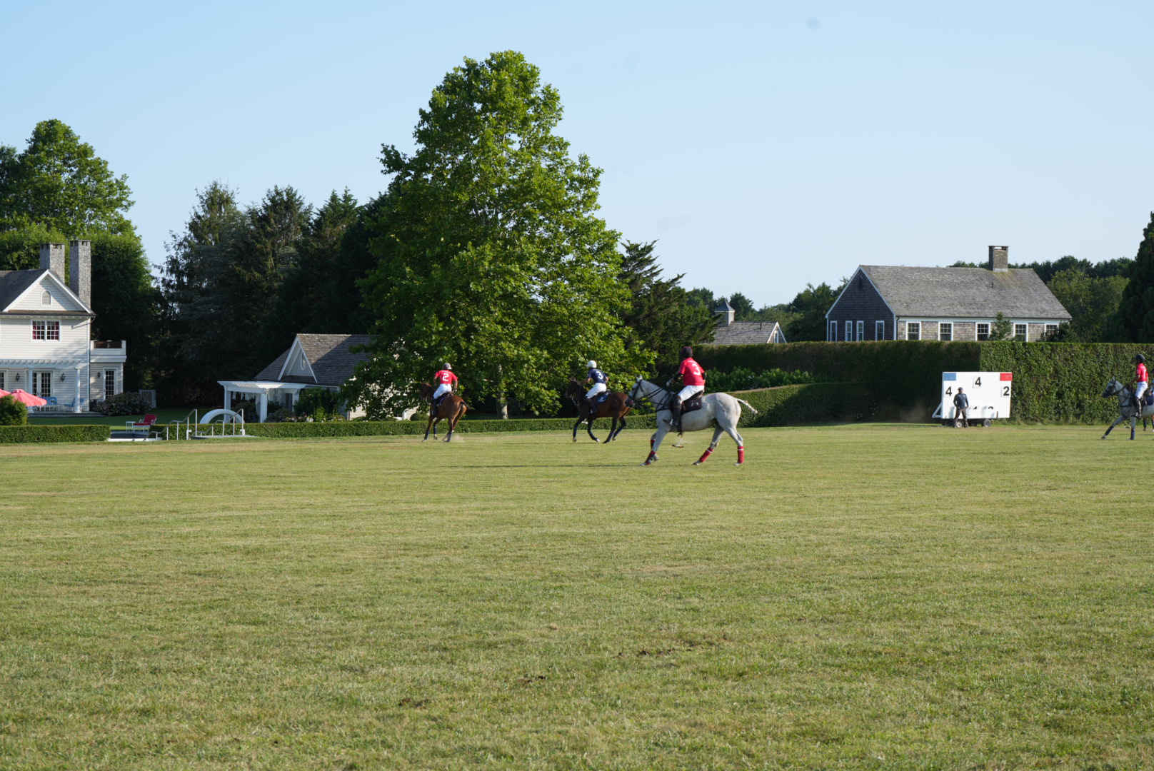 Wide view of the polo field with players riding past shingled Hamptons estates and the scoreboard