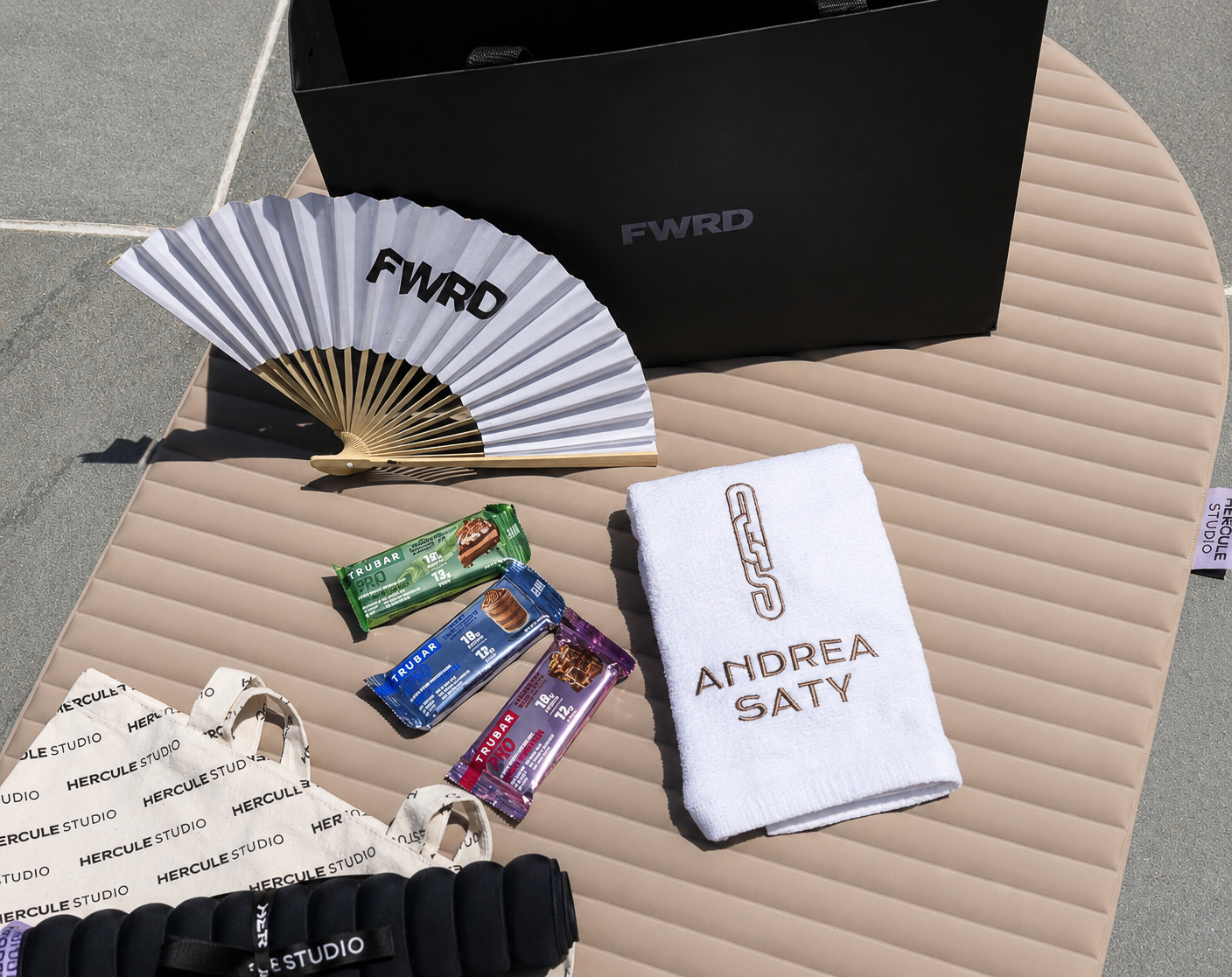 Flat lay of the branded gifting set with FWRD fan, Andrea Saty towel, Trubar bars and Hercule Studio tote