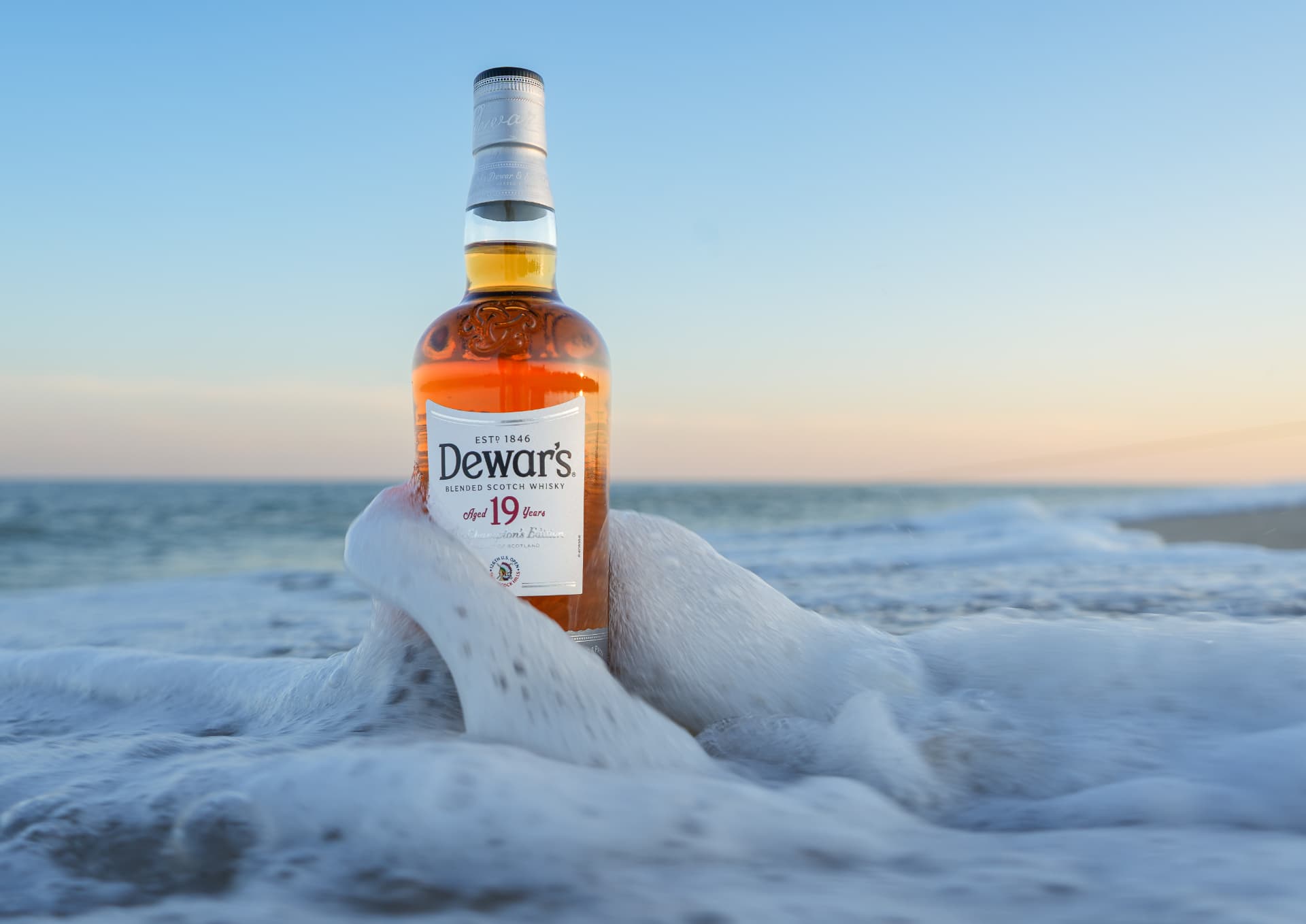Dewar's 19 Year blended scotch whisky standing in ocean surf at sunset