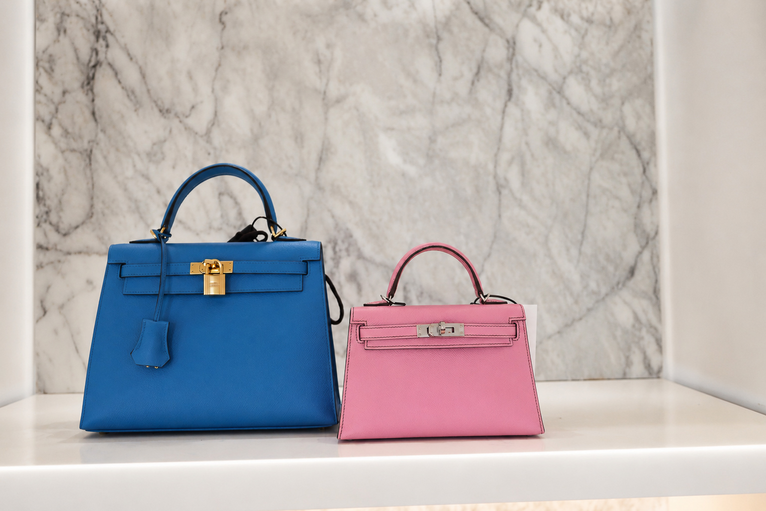 Blue and pink Hermès Kelly handbags styled on a marble display shelf