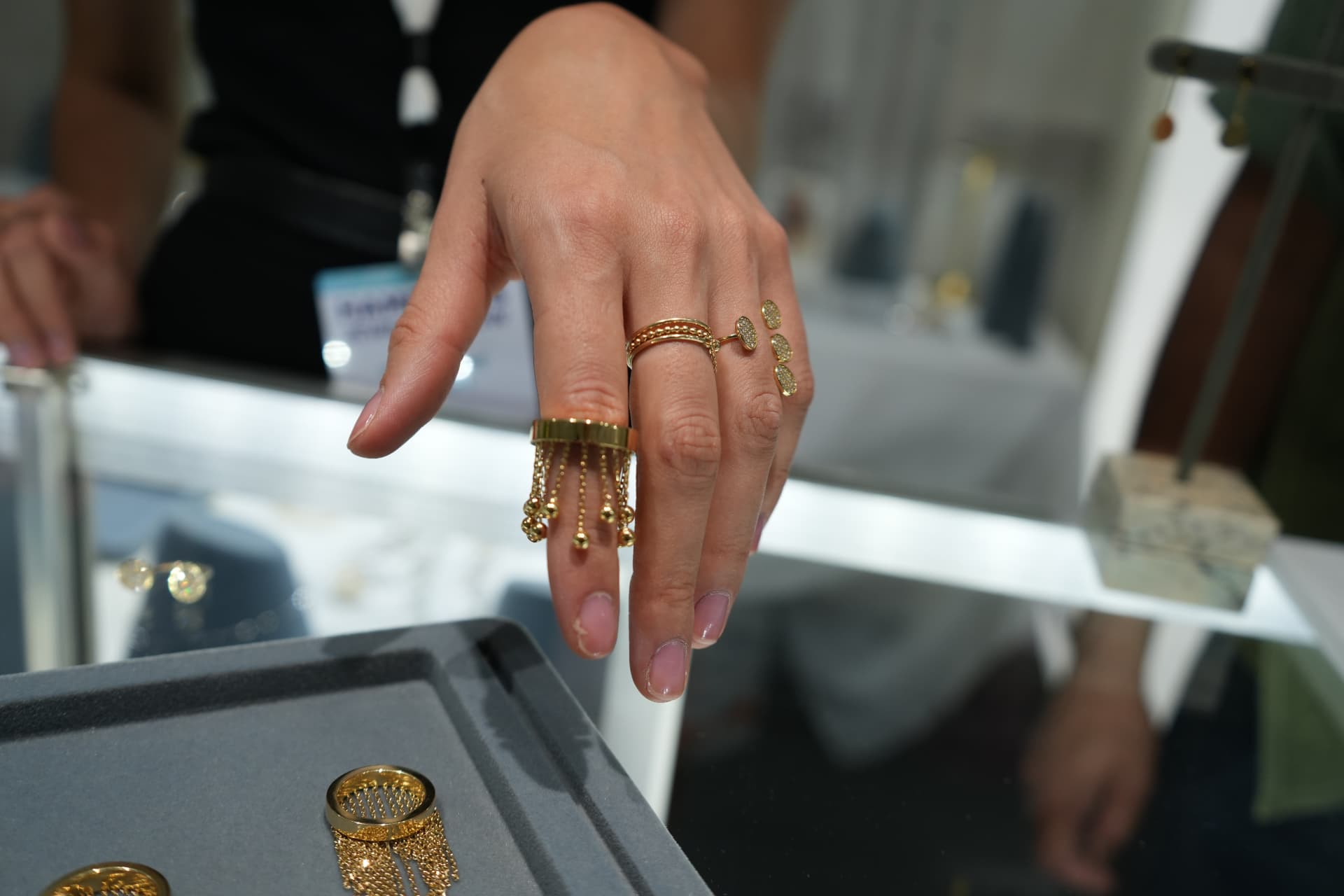 Model's hand displaying a gold fringe ring and pavé chain rings above the lit vitrine